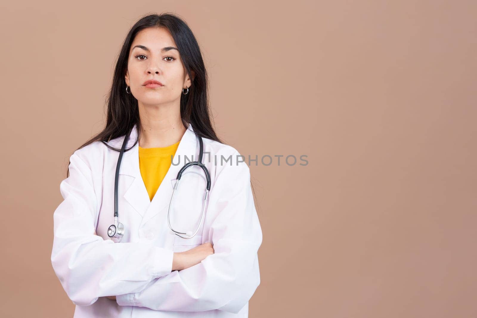Serious hispanic female doctor looking at the camera with arms crossed in studio with brown background
