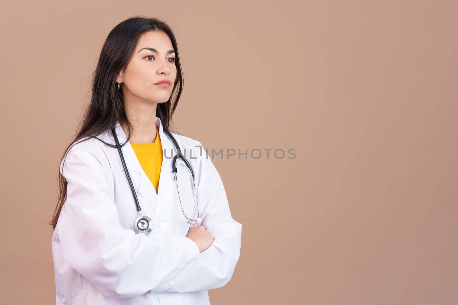 Serious hispanic female doctor with arms crossed looking away in studio with brown background