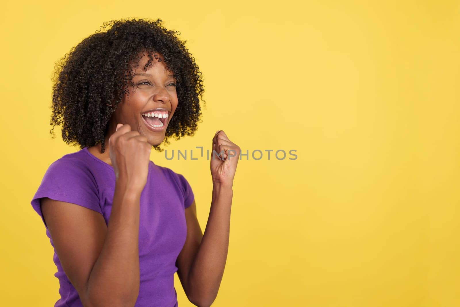 Happy woman with afro hair celebrating while raising fists in studio with yellow background