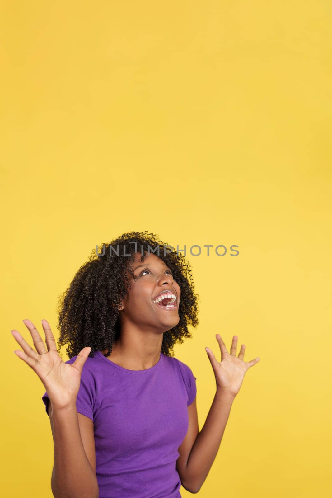 Cool woman with afro hair gesturing happiness looking up in studio with yellow background