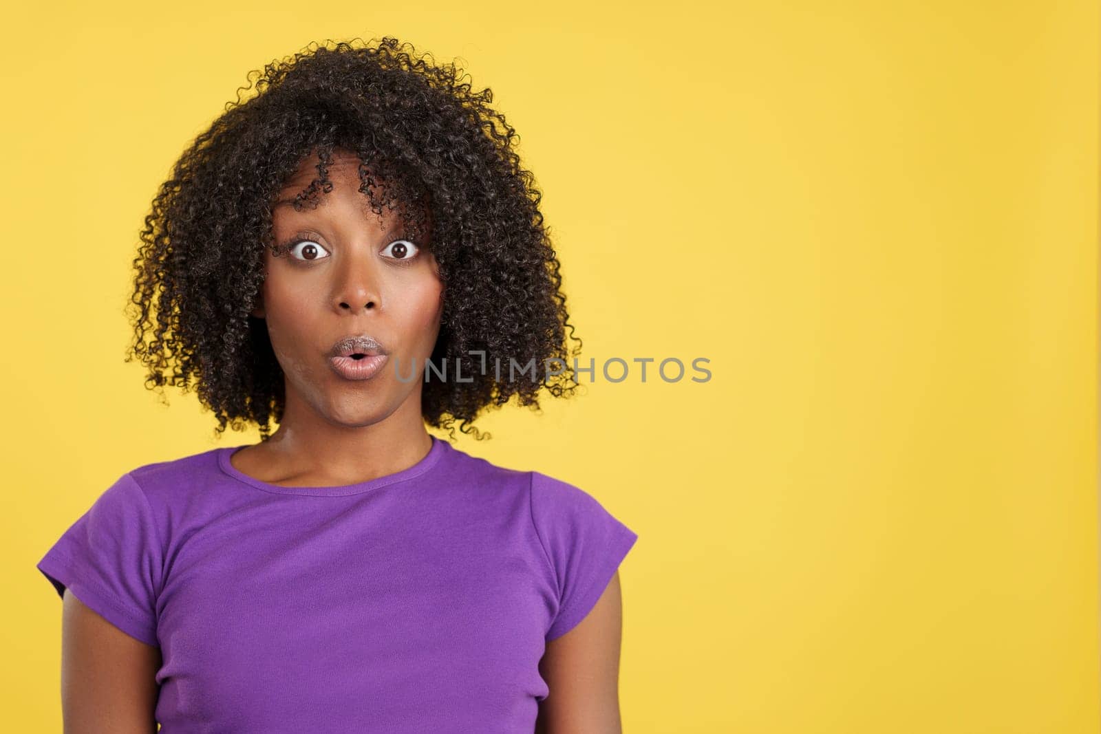 Shocked woman with afro hair looking at camera in studio with yellow background