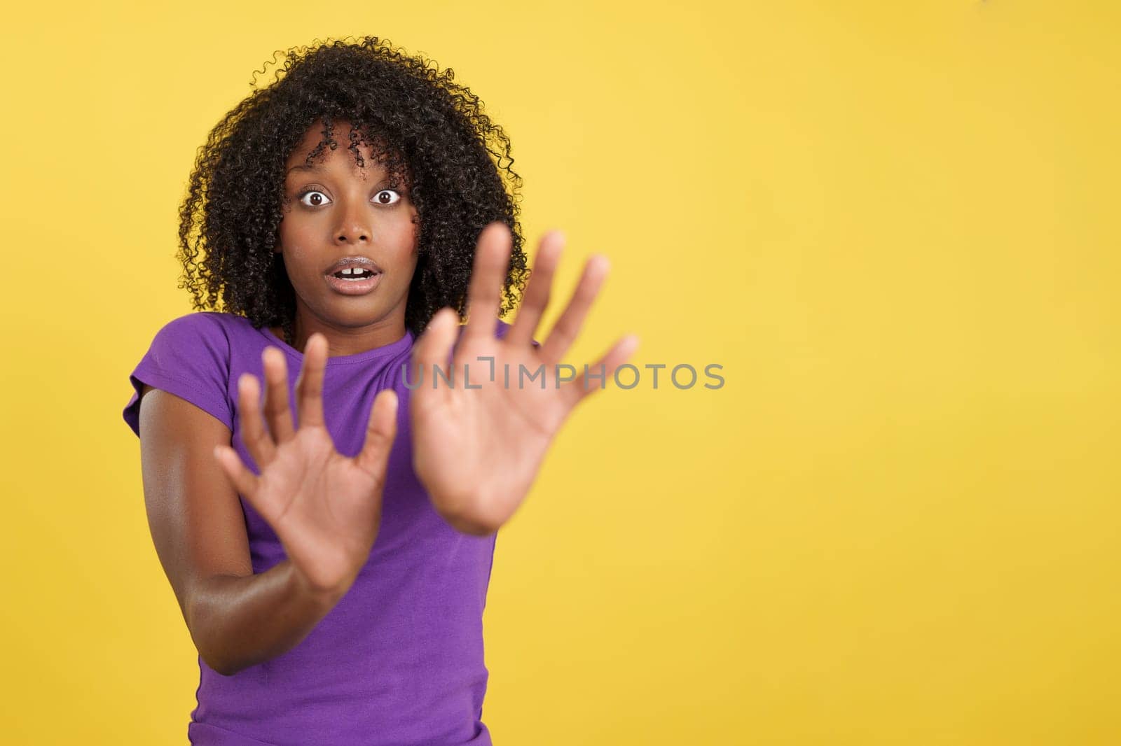 Woman with African hair expressing fear while shielding herself with her hands in studio with yellow background
