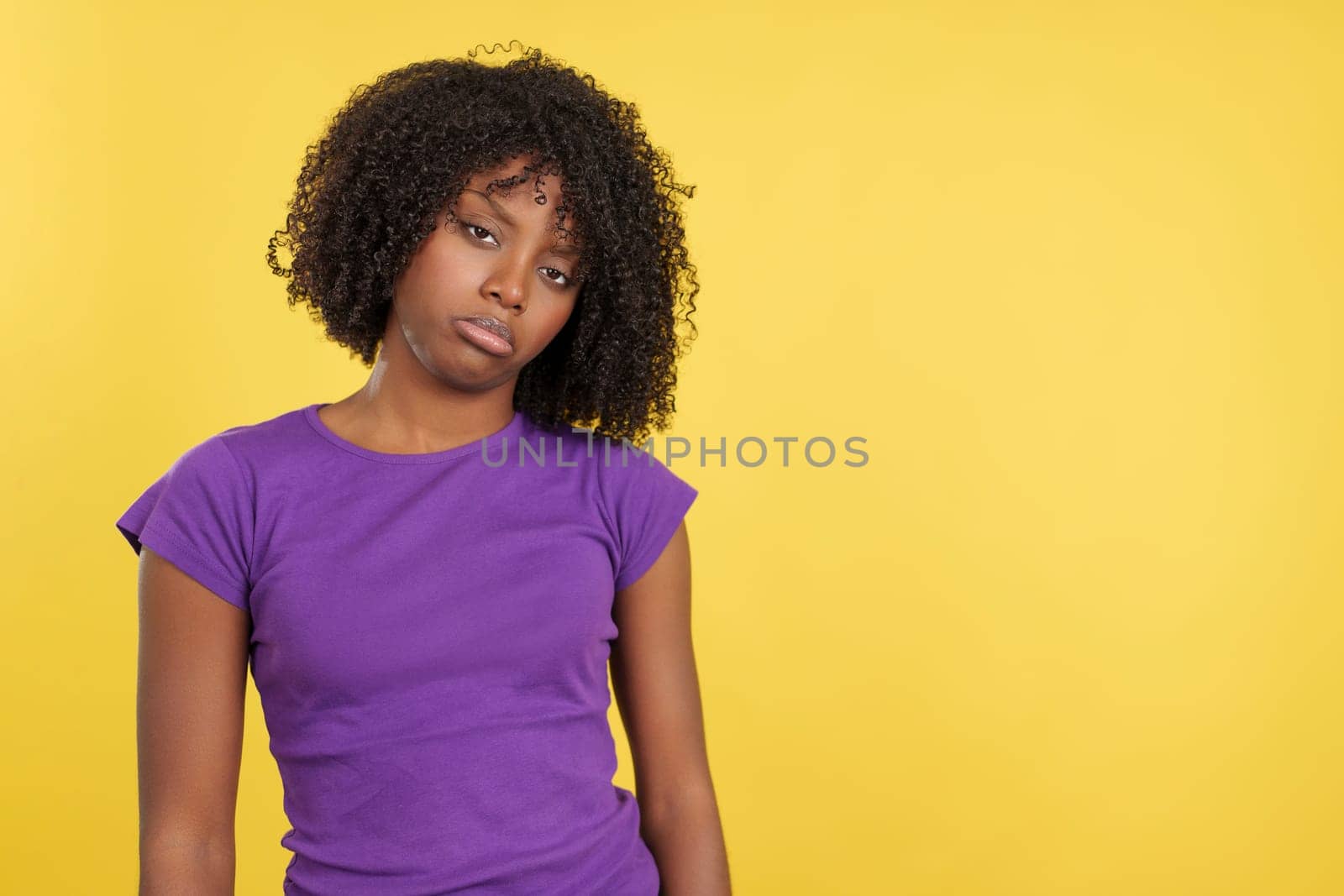 Sad woman with afro hair standing looking at camera in studio with yellow background