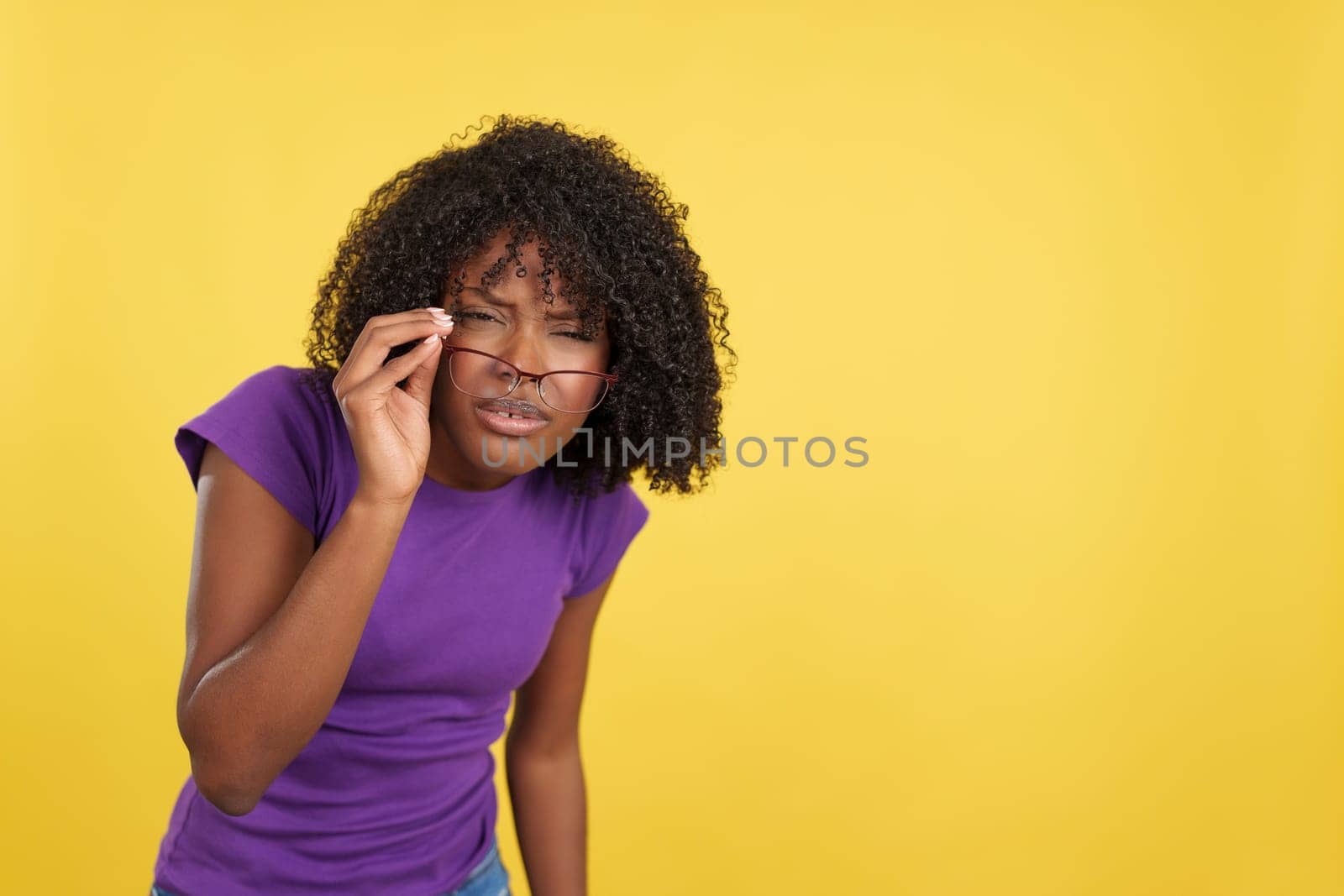 Afro woman taking off her glasses to look at the camera in studio with yellow background