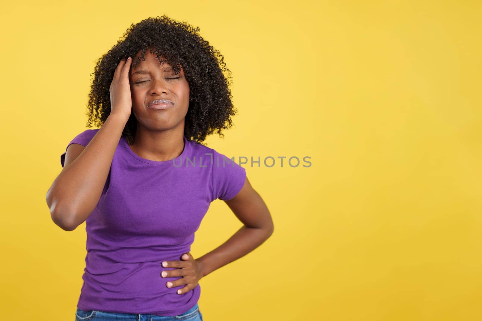 Woman with afro hair standing while gesturing headache in studio with yellow background