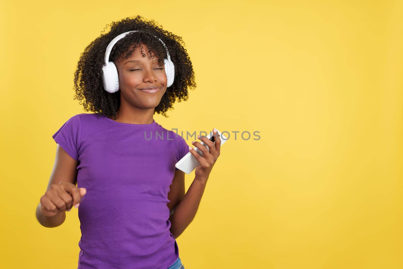 Cool woman with afro hair listening to music with headphones and mobile in studio with yellow background