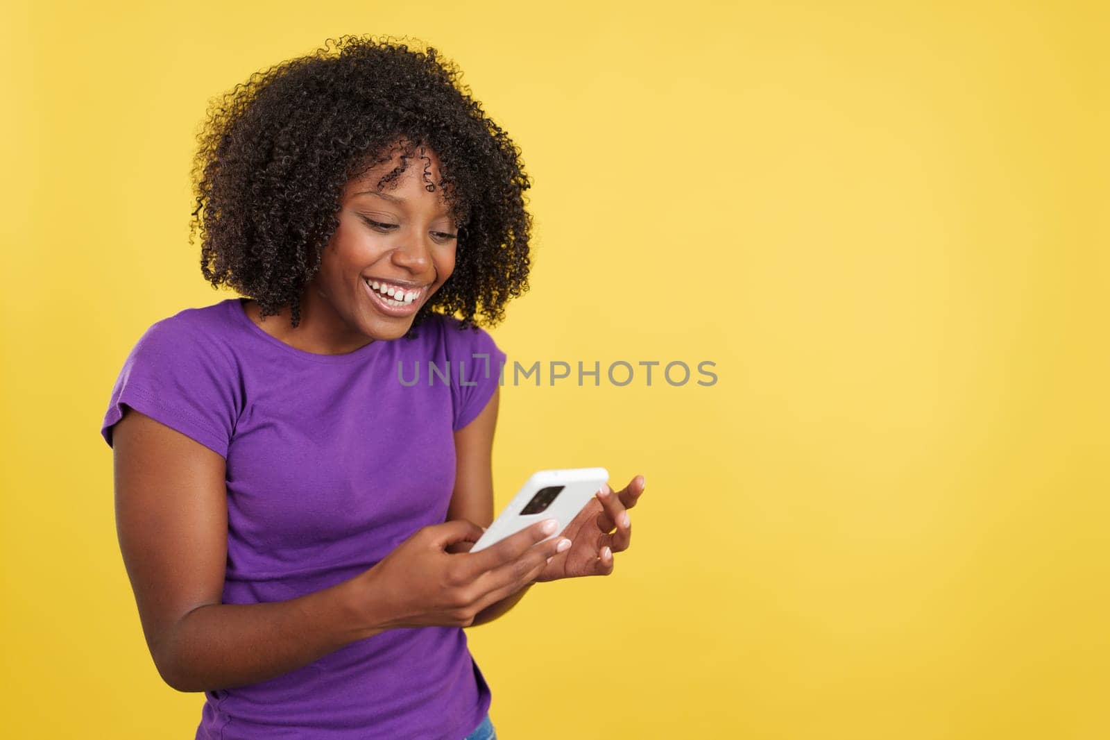 Happy woman with afro hair smiling while reading a mobile in studio with yellow background
