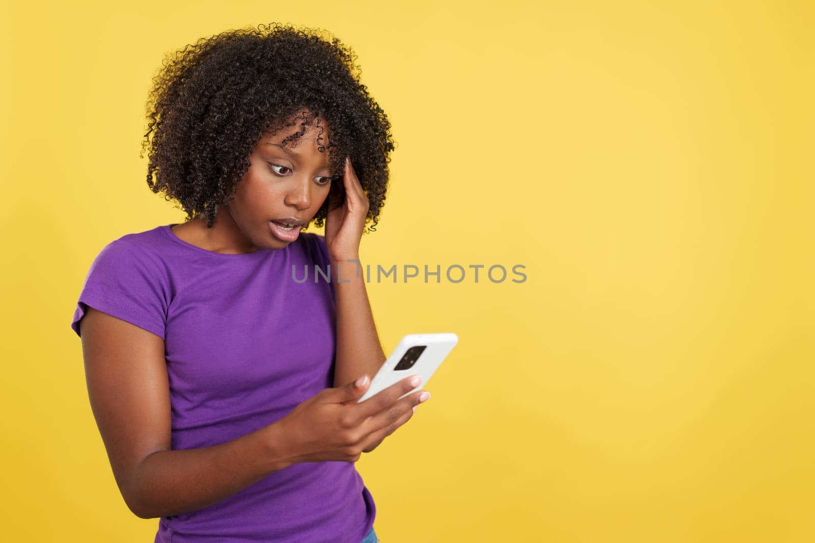 Worried woman with afro hair using a mobile phone in studio with yellow background