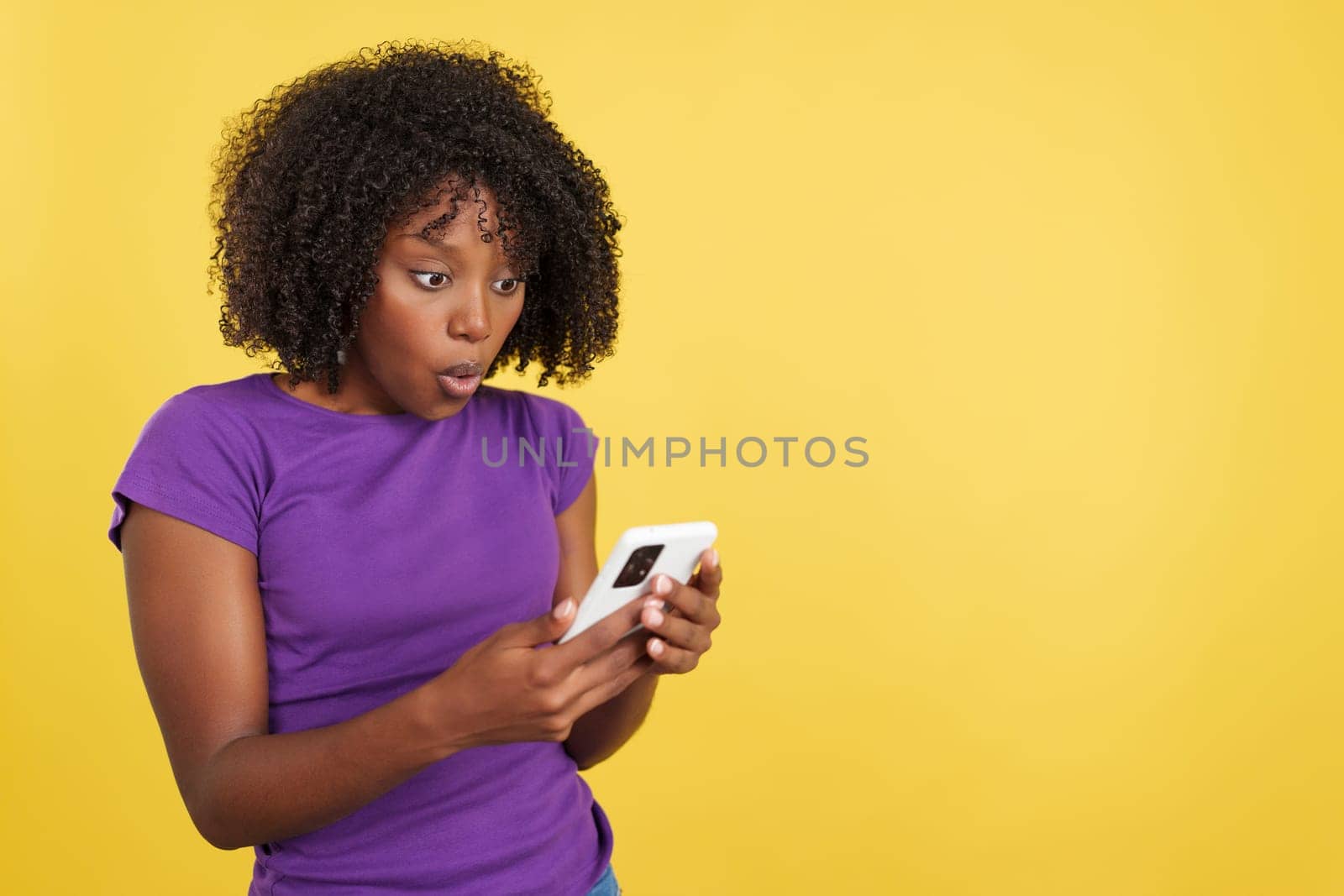Surprised woman with afro hair using a mobile in studio with yellow background