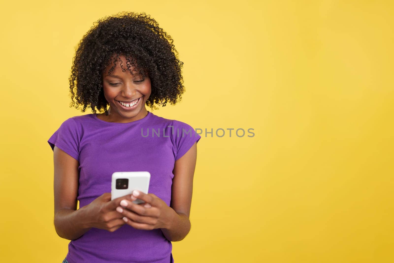 Woman with afro hair smiling while sending a message with a mobile in studio with yellow background