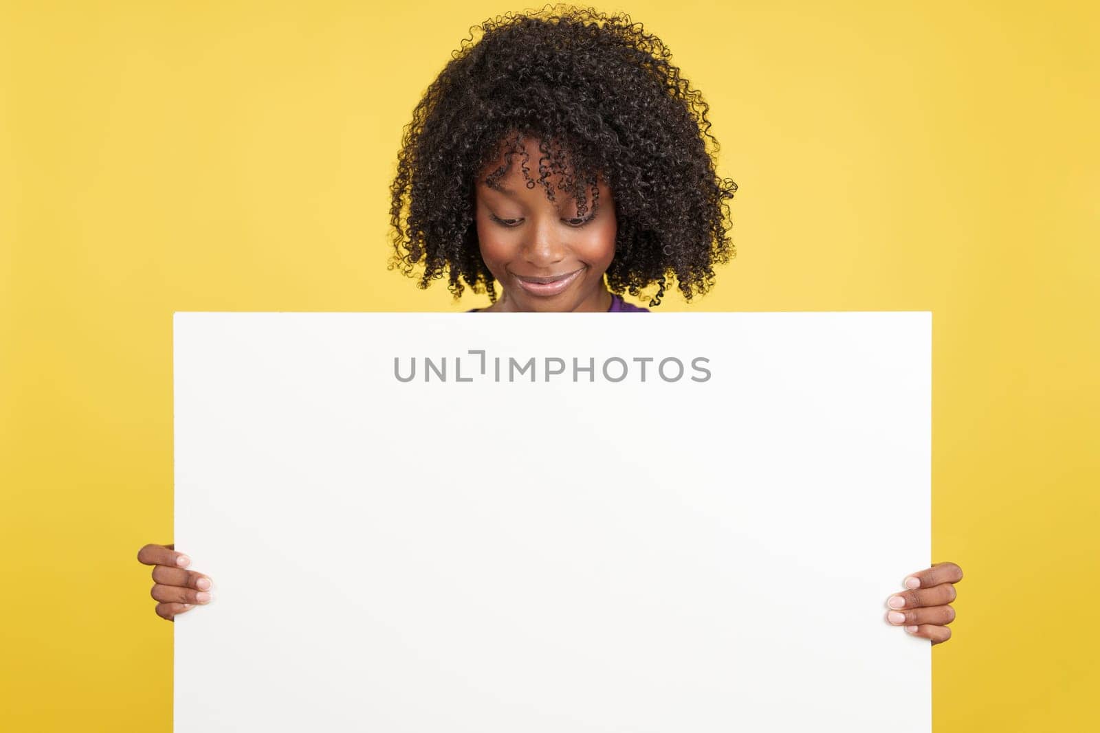 Woman with afro hair looking down to a blank board that she is holding in studio with yellow background