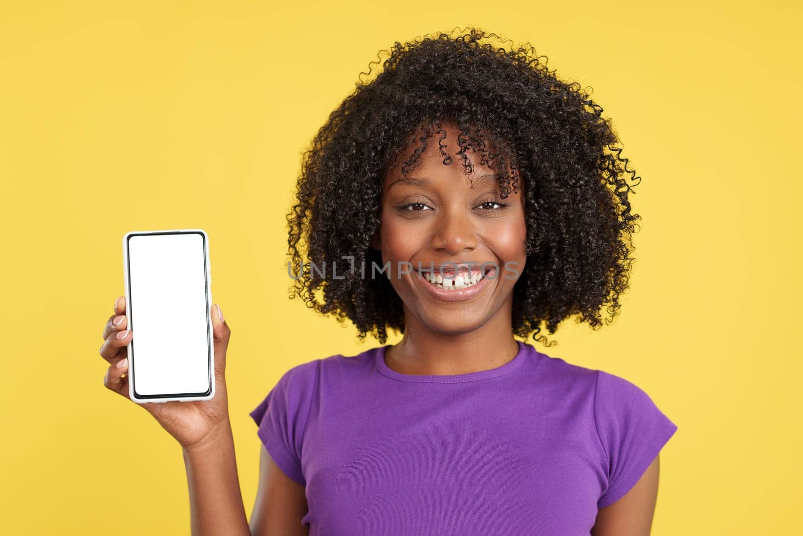 Woman with afro hair smiling at the camera holding a mobile with a white screen in studio with yellow background