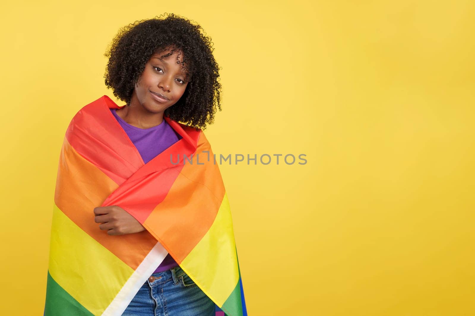 Woman with afro hair wrapped with a lgbt rainbow flag in studio with yellow background