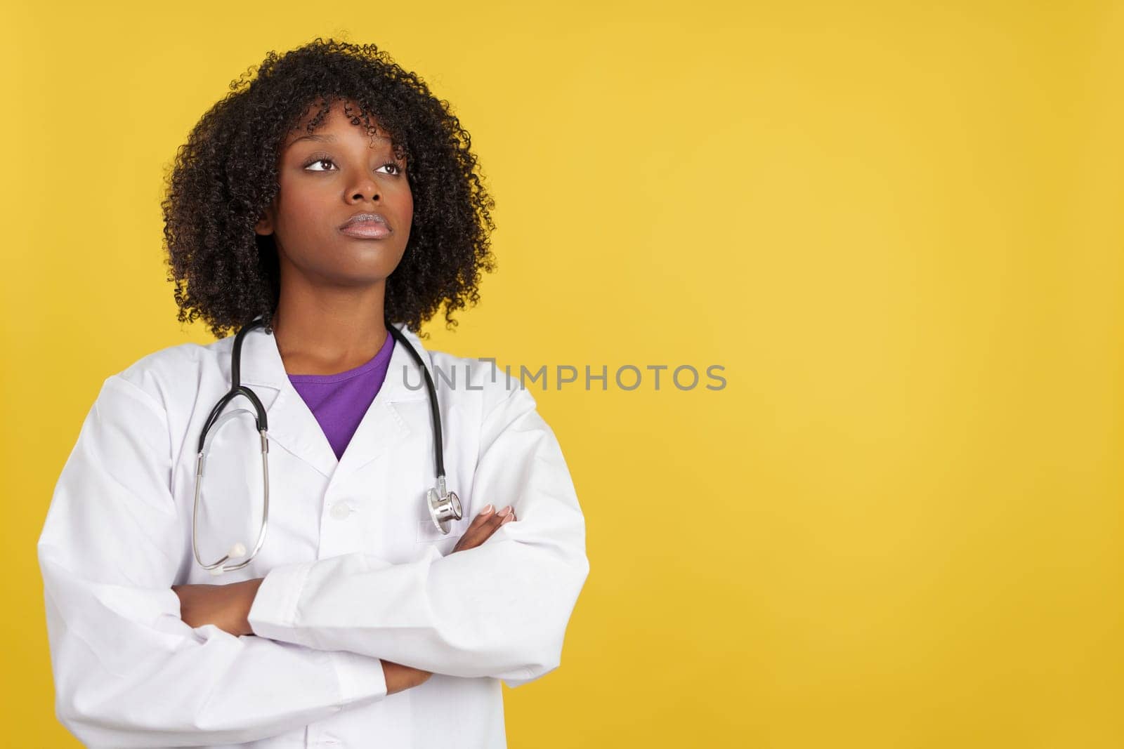 Proud afro female doctor looking up with arms crossed in studio with yellow background