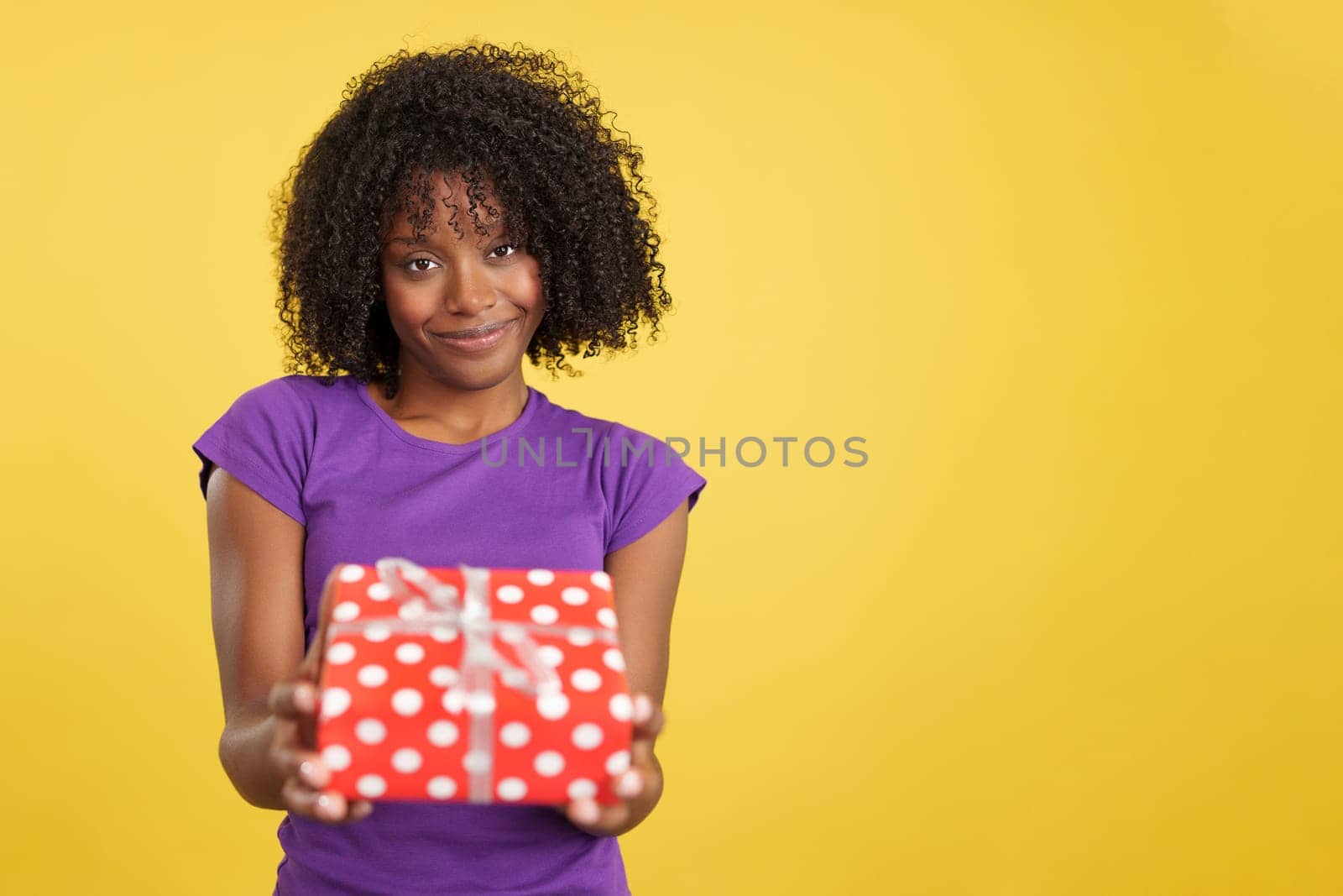 Generous woman with afro hair giving a present while looking at camera in studio with yellow background