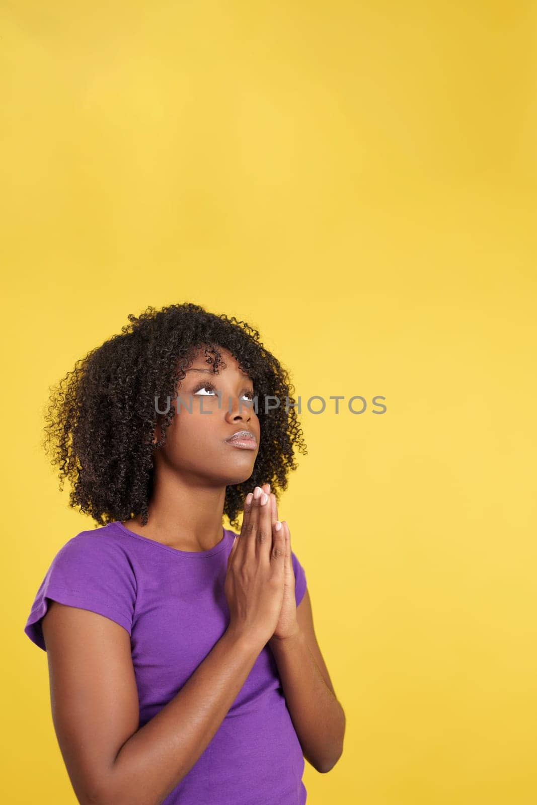 Afro woman praying while looking up joining hands in studio with yellow background