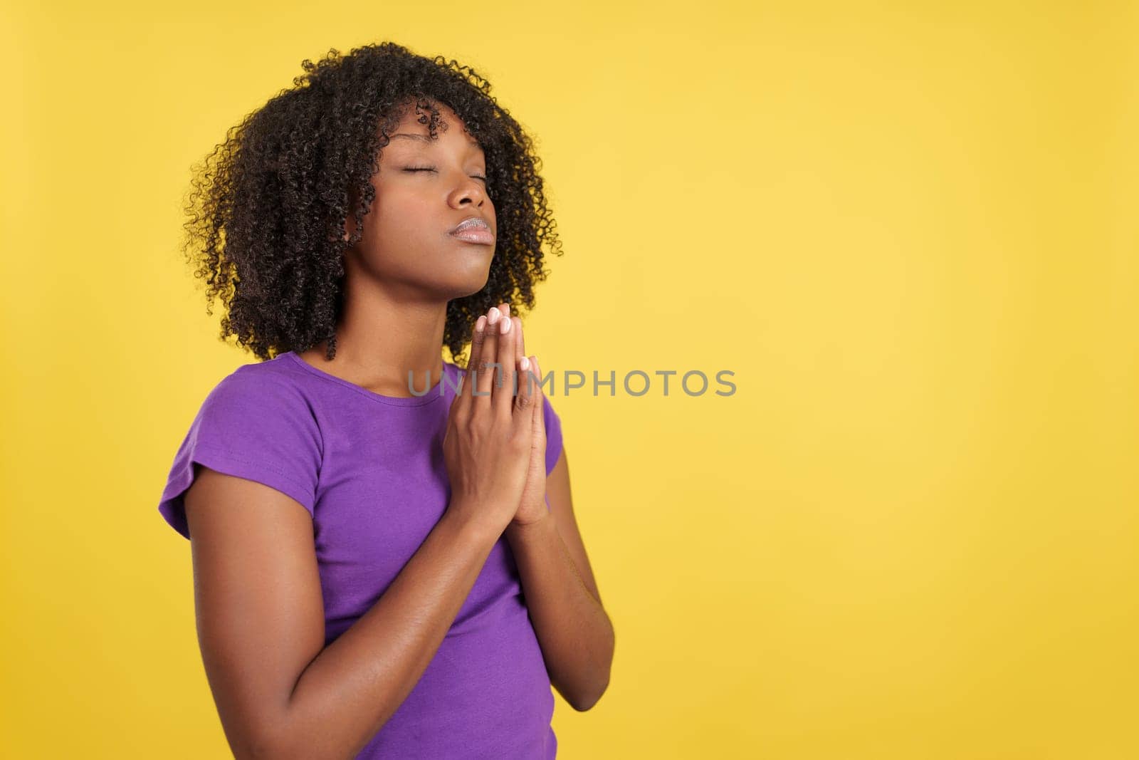 Woman with afro hair praying looking up with eyes closed and folded hands in studio with yellow background