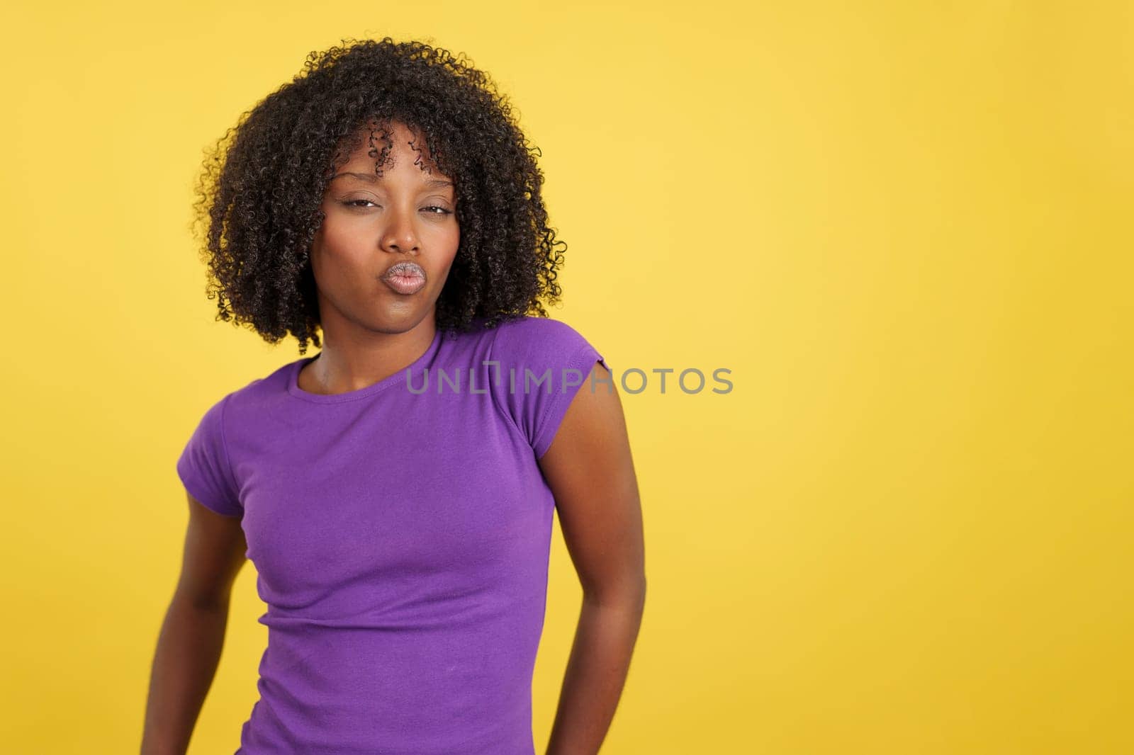 Provocative woman with afro hair kissing at camera in studio with yellow background