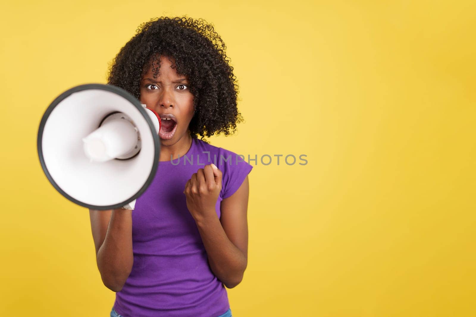 Angry African woman shouting using a megaphone and gesturing with fist in studio with yellow background