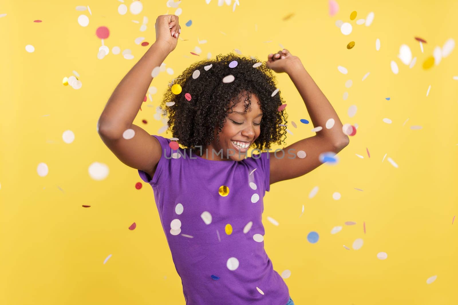 Cool Afro woman dancing and celebrating surrounded by confetti int he air in studio with yellow background