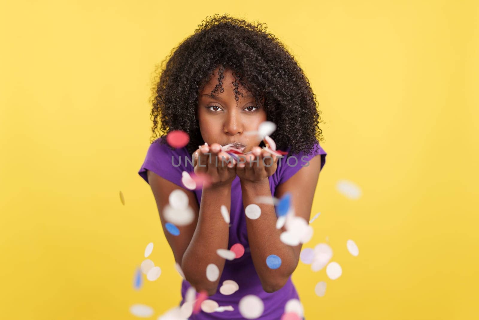 Cheerful Afro woman blowing confetti while celebrate in studio with yellow background