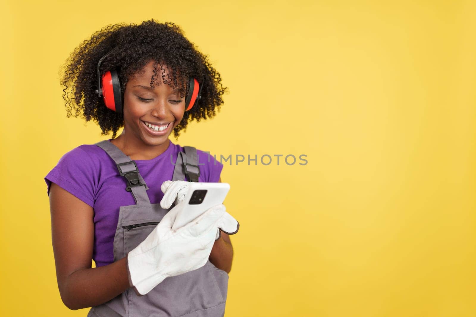 Afro female carpenter standing using the mobile in studio with yellow background