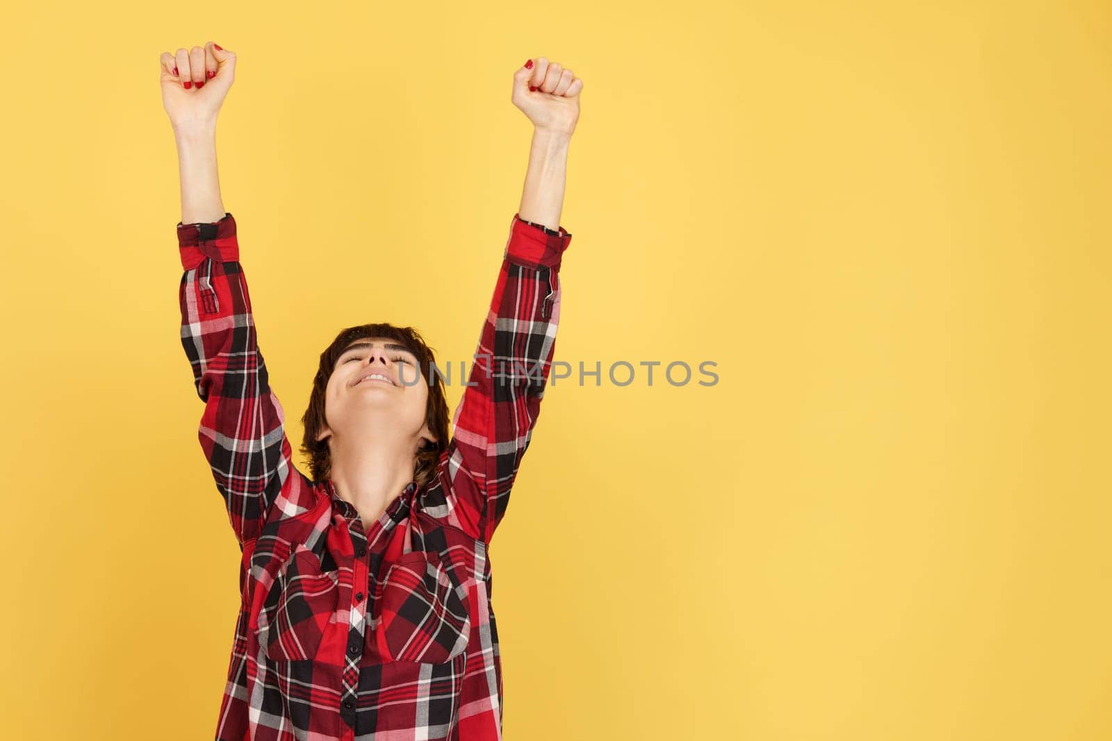 Happy androgynous person celebrating with arms up in studio with yellow background