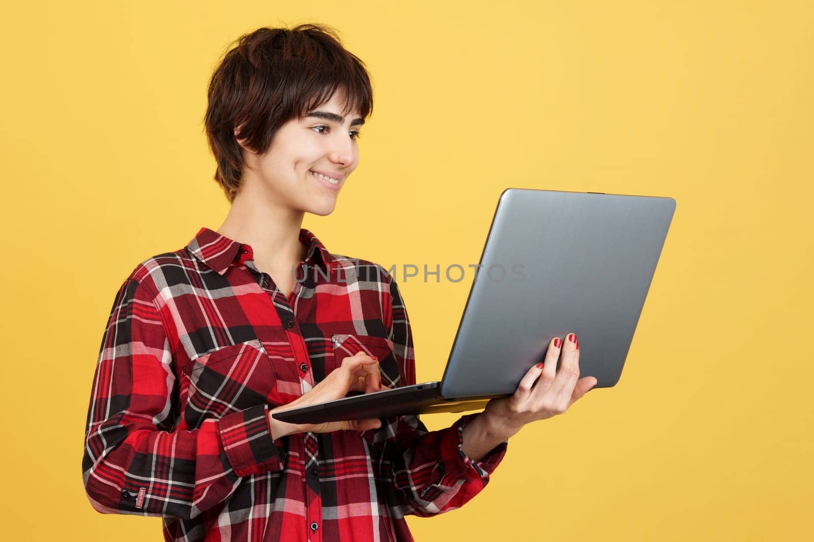 Androgynous person smiling while using a laptop in studio with yellow background