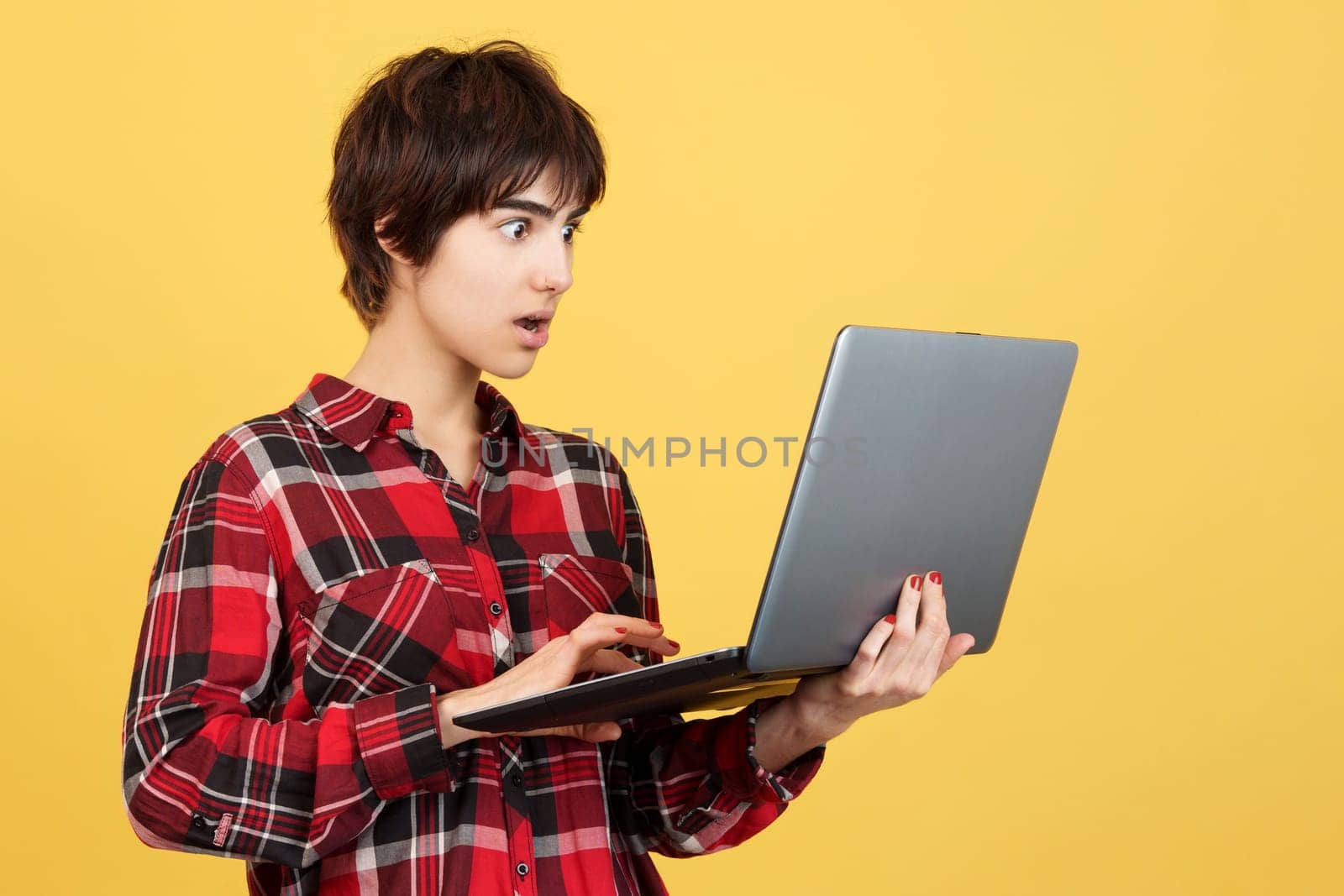 Surprised androgynous person standing and using a laptop in studio with yellow background