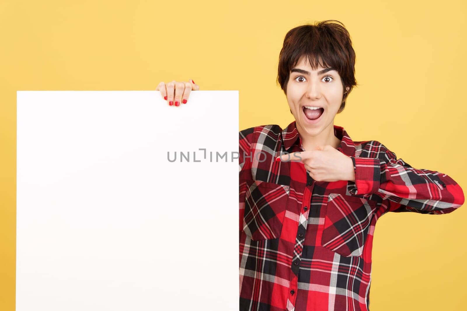 Amazed androgynous person pointing a blank panel in studio with yellow background