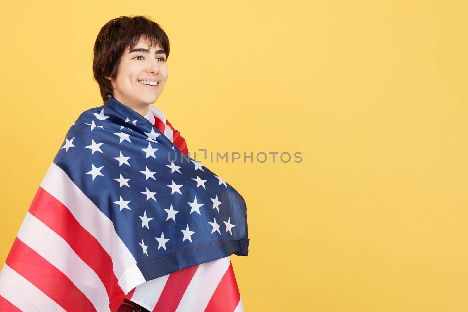 Androgynous person wrapped with a USA flag in studio with yellow background