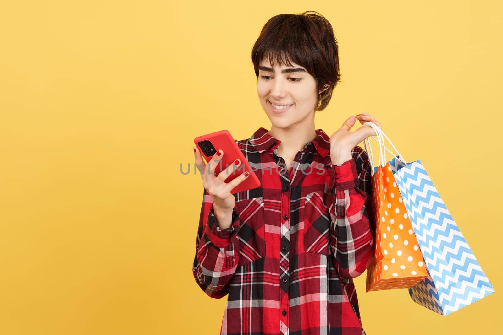Androgynous person using a mobile while holding shopping bags in studio with yellow background