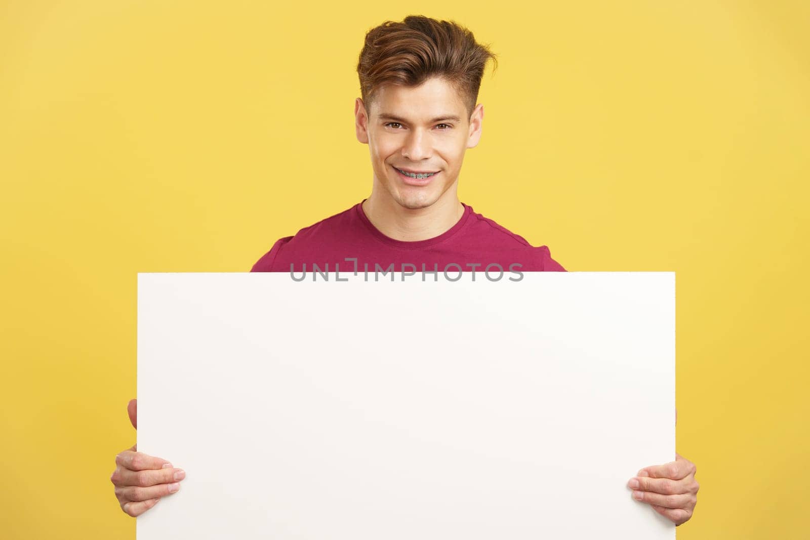 Caucasian man holding a blank panel smiling at the camera in studio with yellow background
