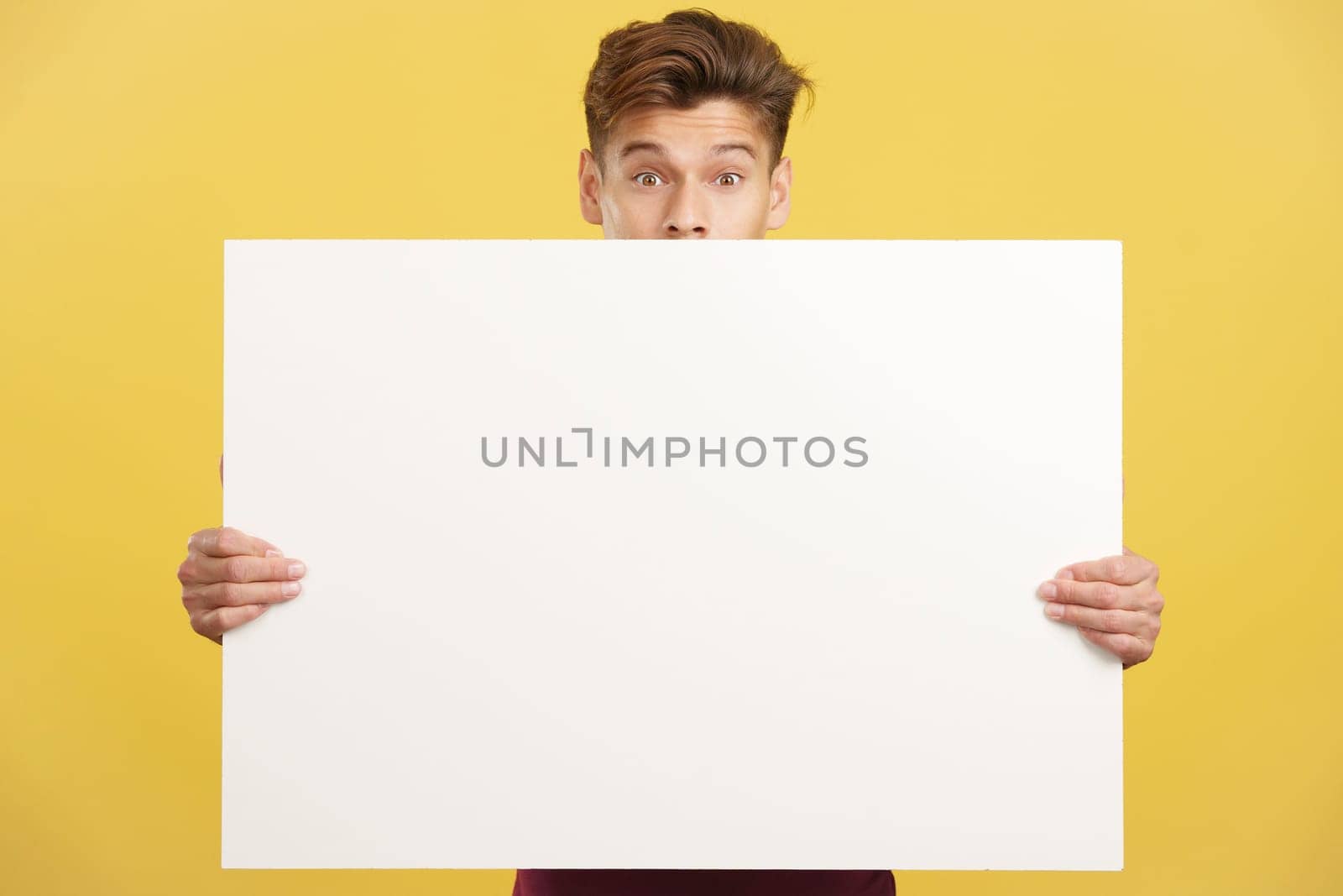 Surprised man hiding behind a blank panel in studio with yellow background