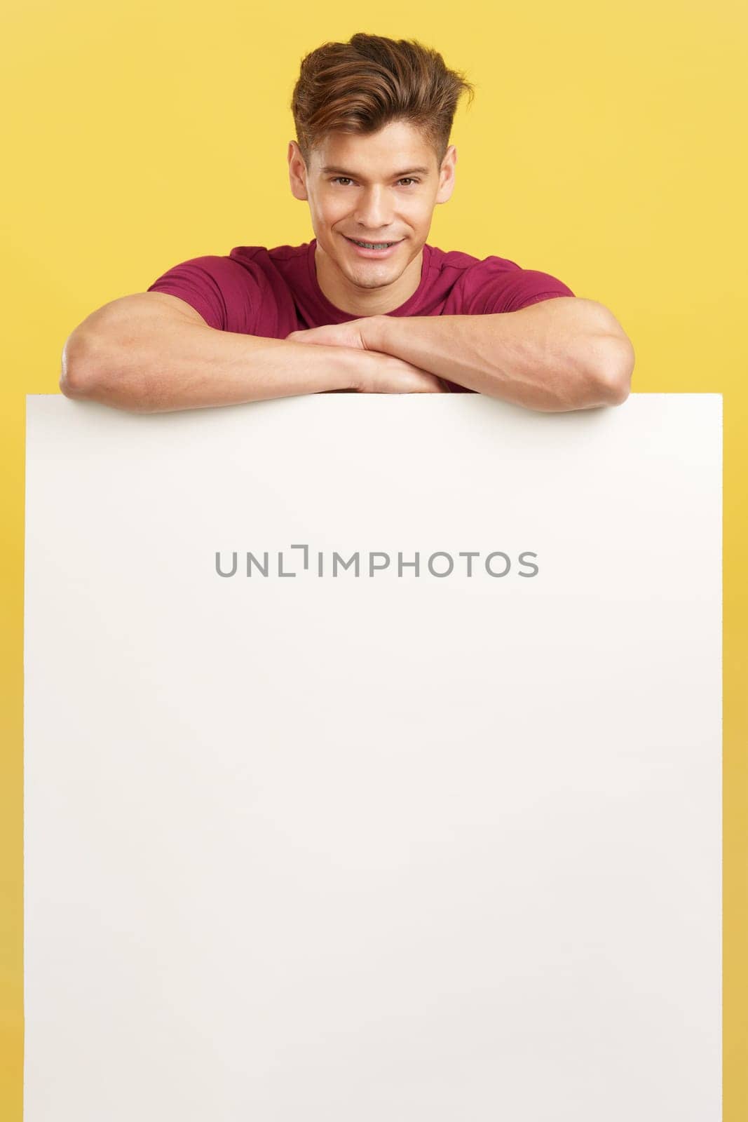 Blonde handsome man leaning on a blank panel in studio with yellow background