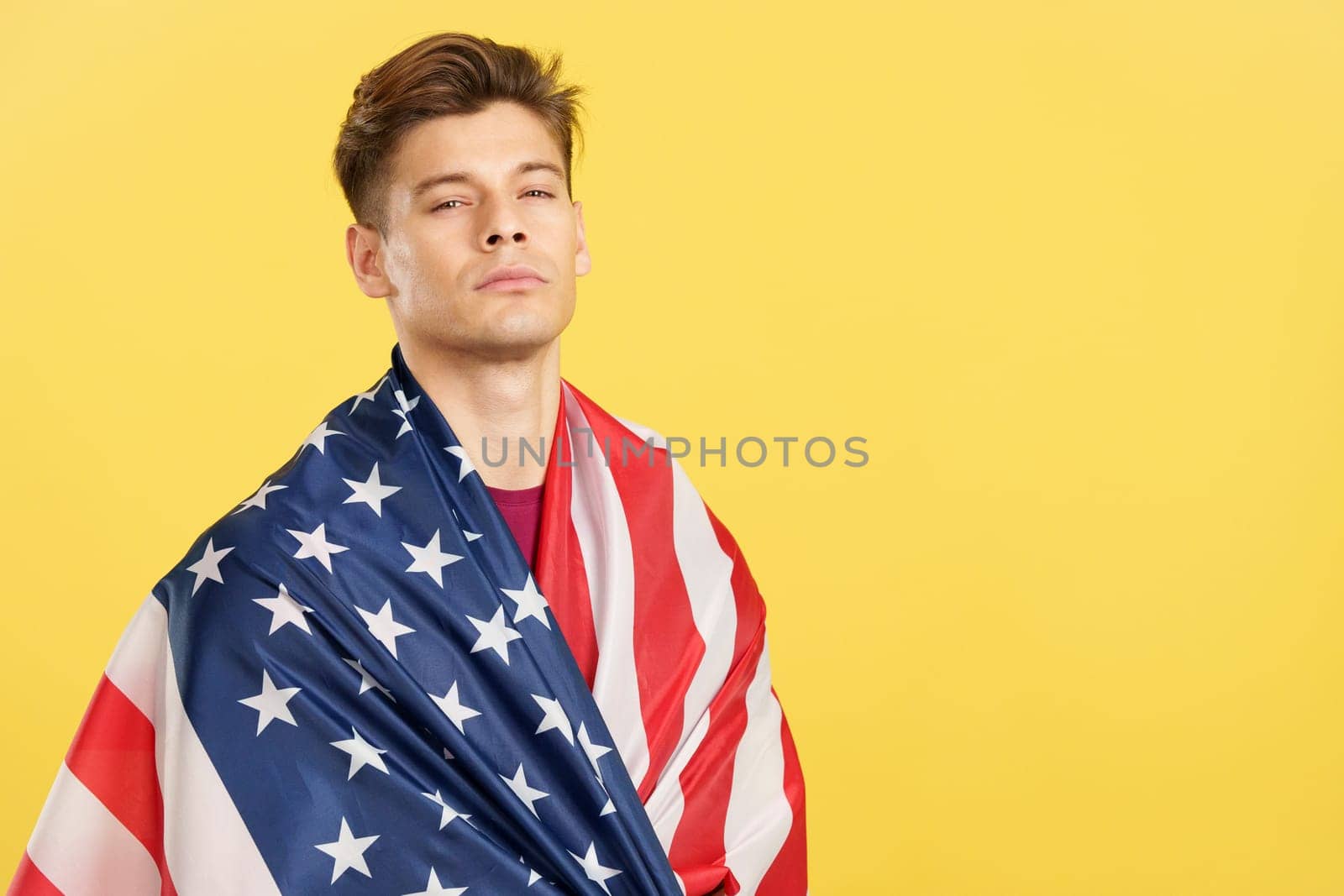 Caucasian man with a look of superiority wrapping with a United States national flag in studio with yellow background