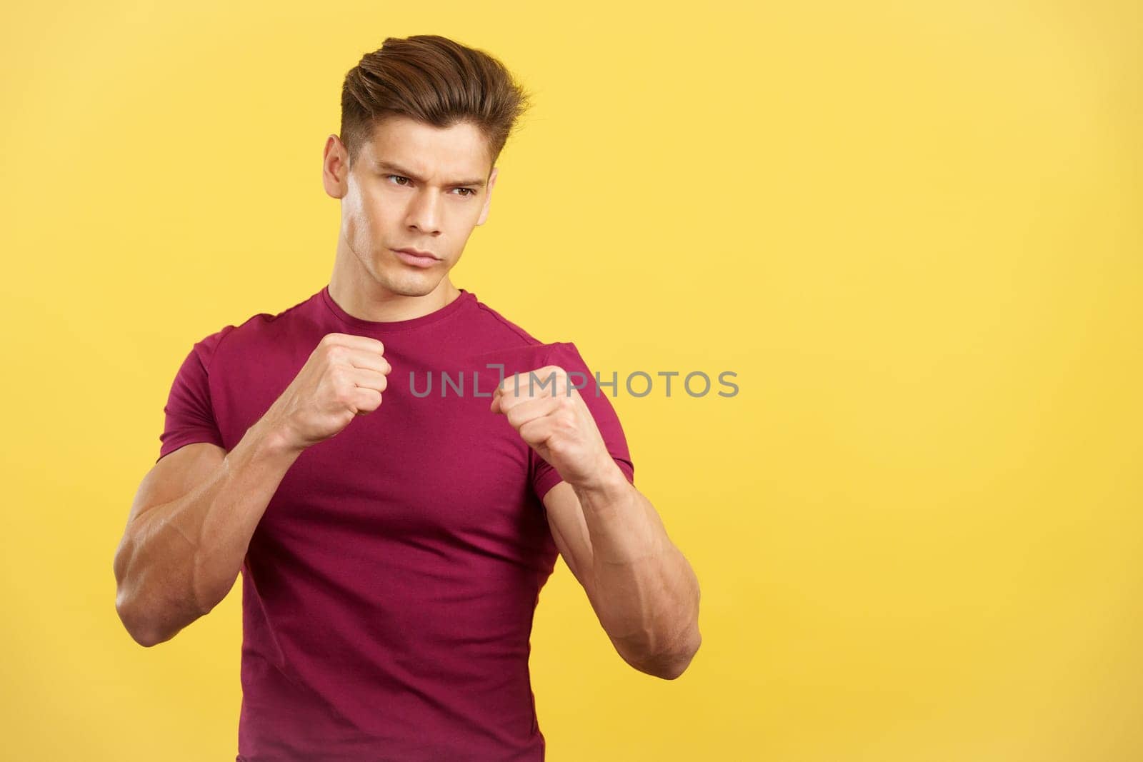 Strong man in combat pose raising fists in boxing guard in studio with yellow background