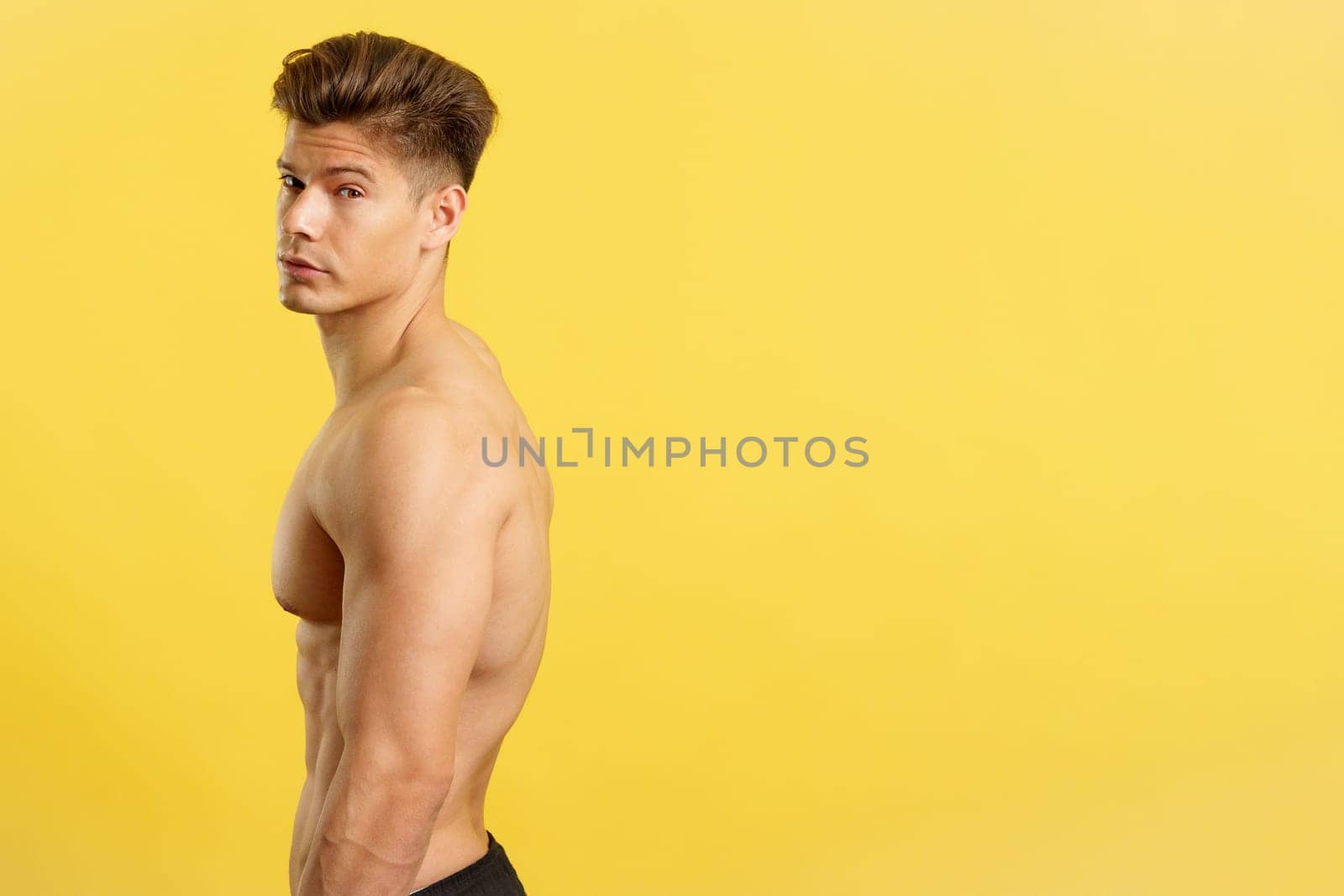 Handsome man with bare torso turning to looking at camera in studio with yellow background