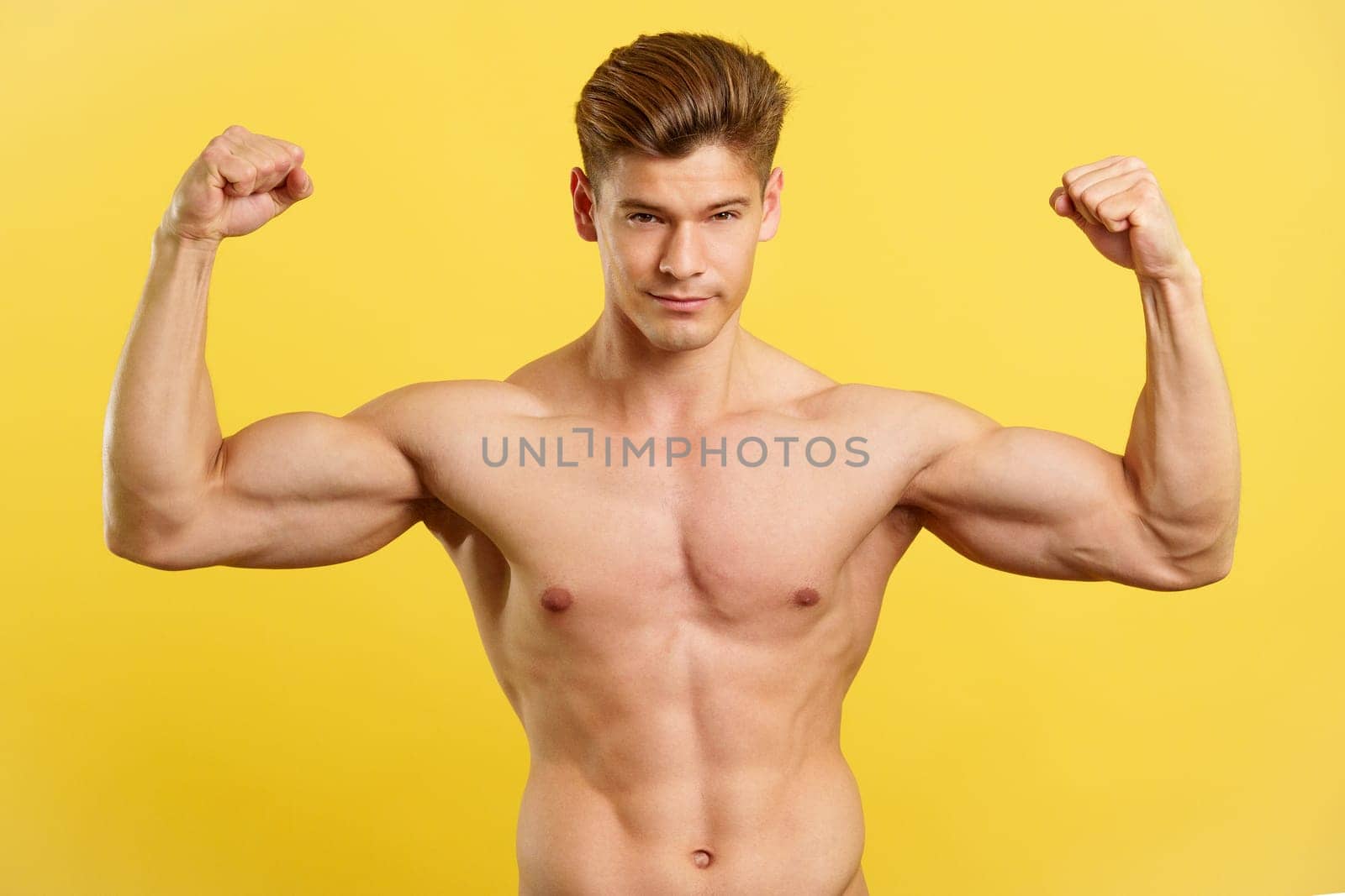Strong man showing biceps while looking at camera with the bare torso in studio with yellow background