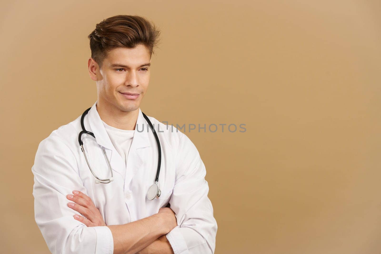 Handsome doctor smiling with arms crossed looking away in studio with brown background