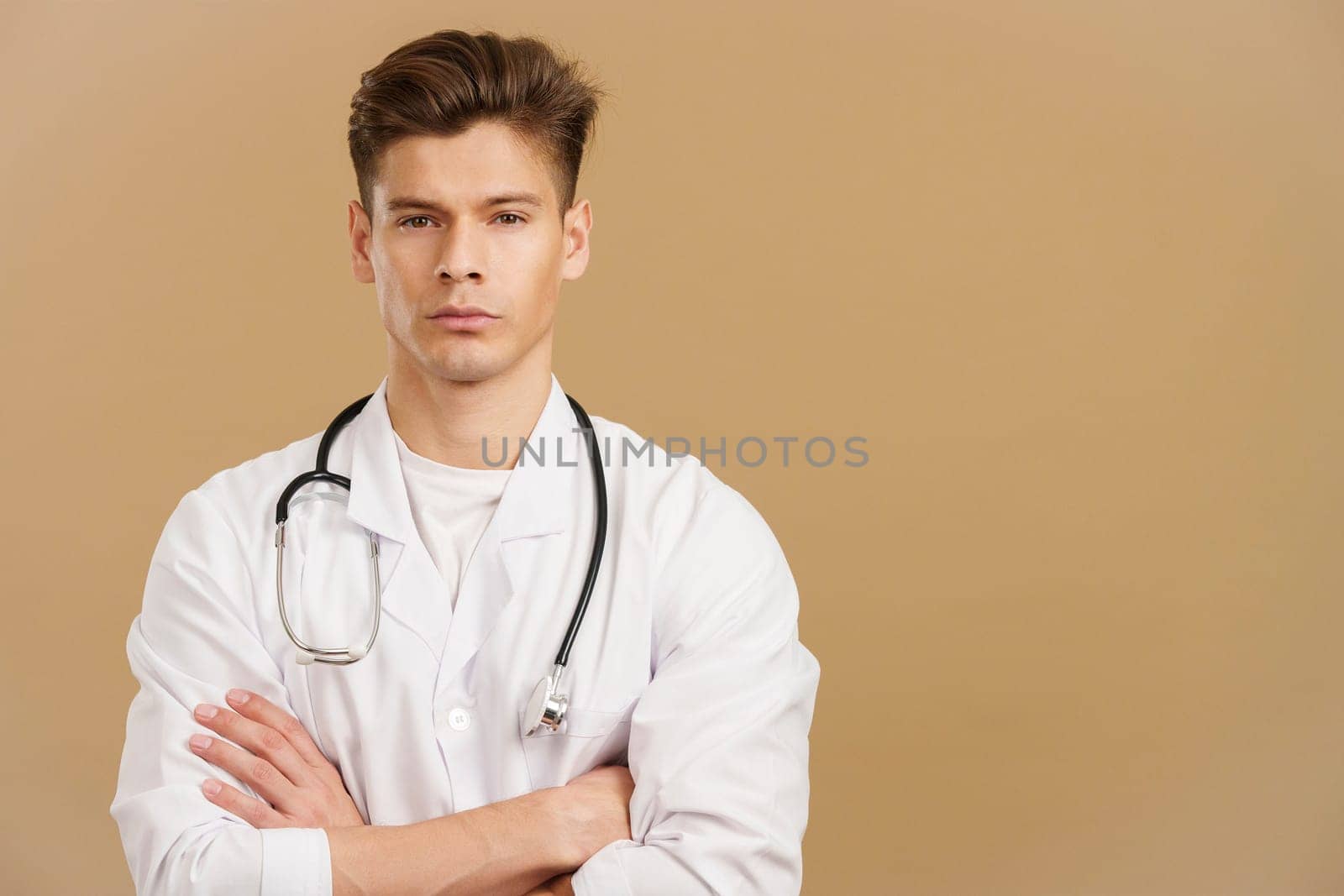 Handsome serious doctor looking at camera standing with arms crossed in studio with brown background