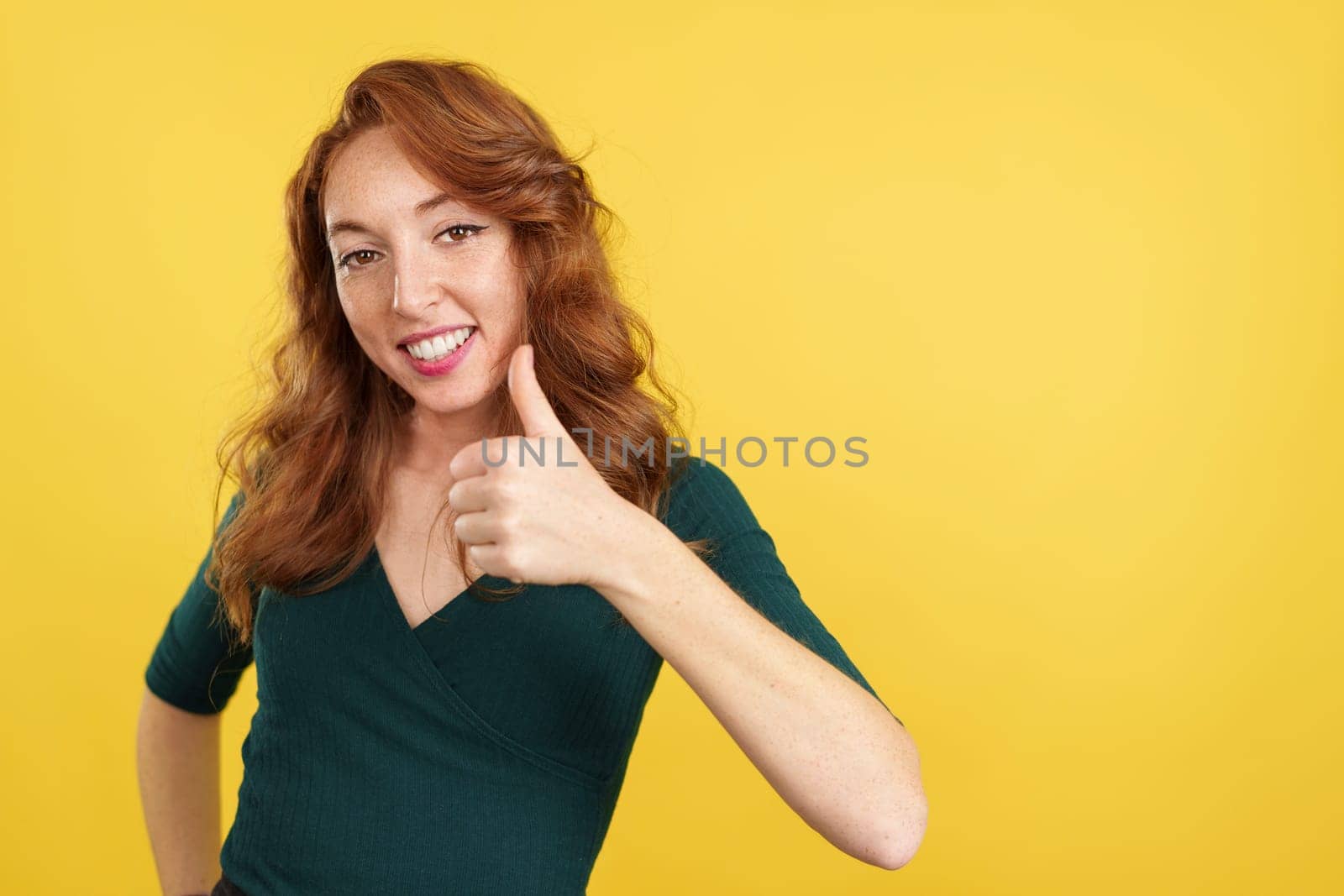 Happy redheaded woman gesturing agreement raising a thumb up in studio with yellow background