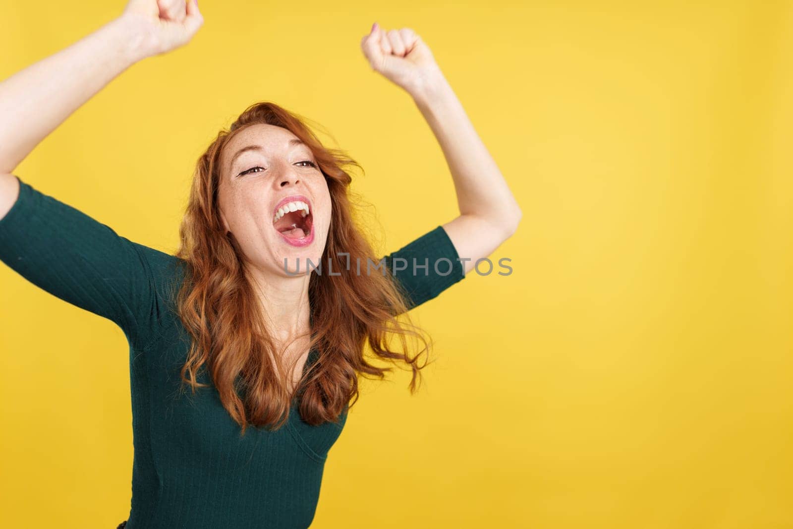 Happy redheaded woman celebrating while raising arms in studio with yellow background