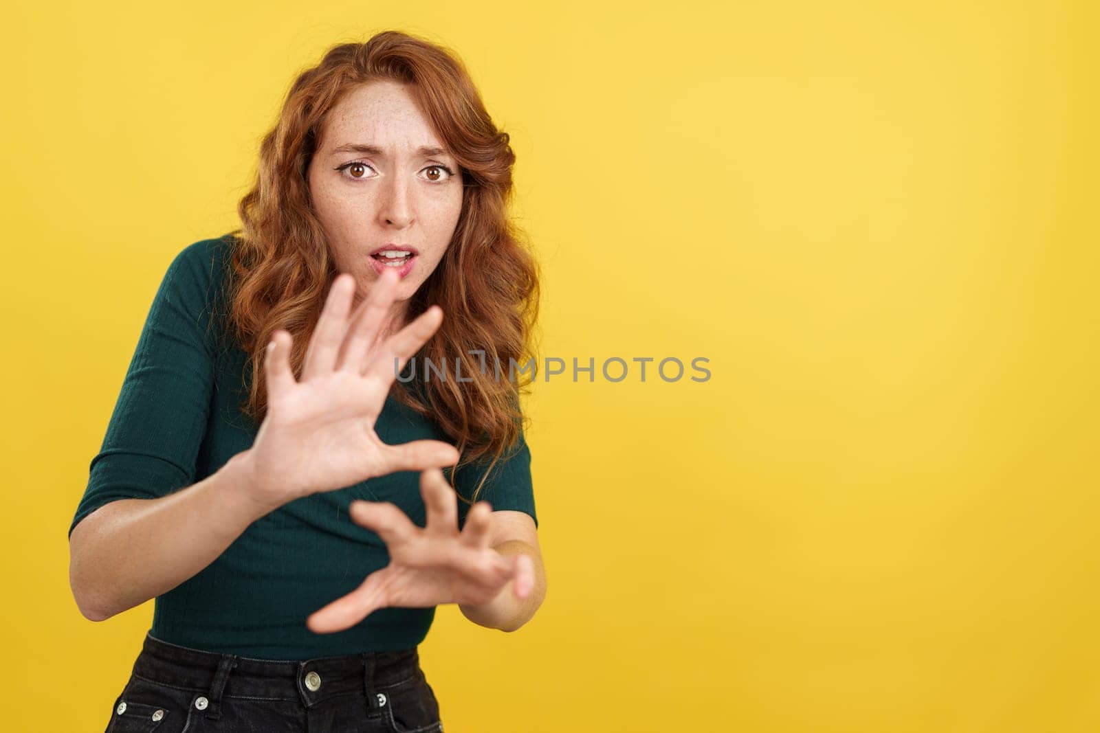 Redheaded woman gesturing with the hands in fear in studio with yellow background