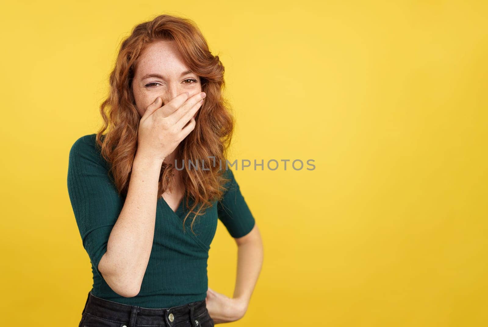 Shy redheaded woman covering the mouth while smiling in studio with yellow background