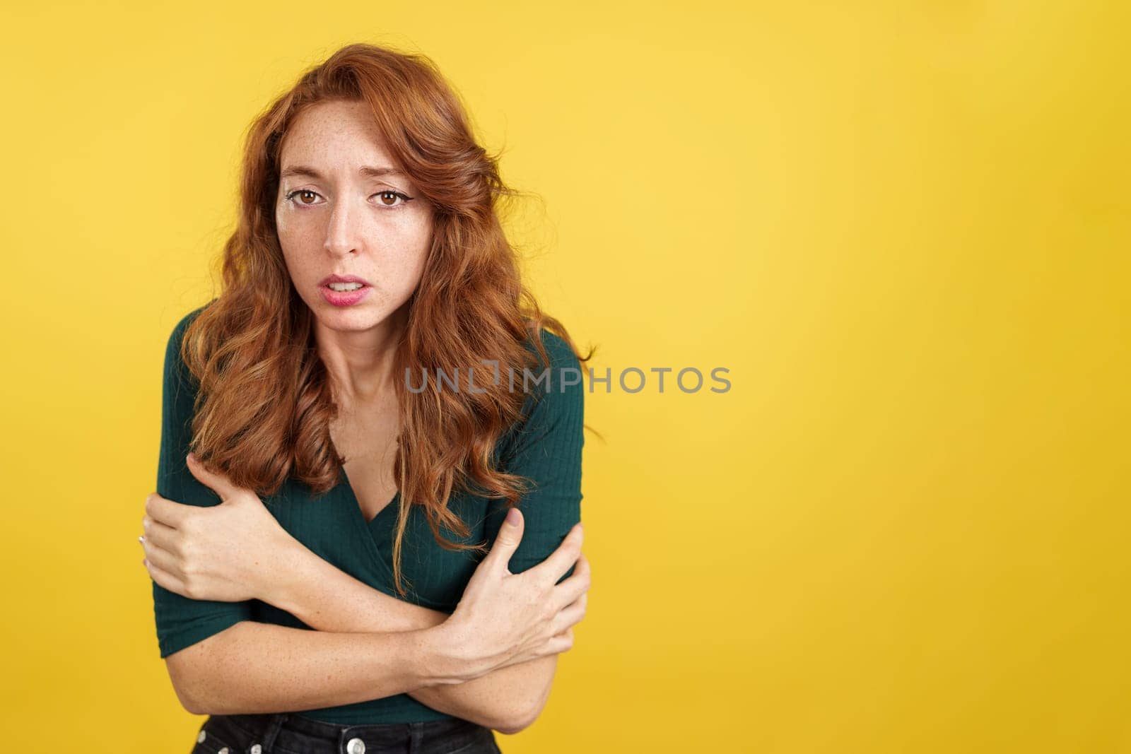 Redheaded woman looking at camera woman gesturing she is cold in studio with yellow background