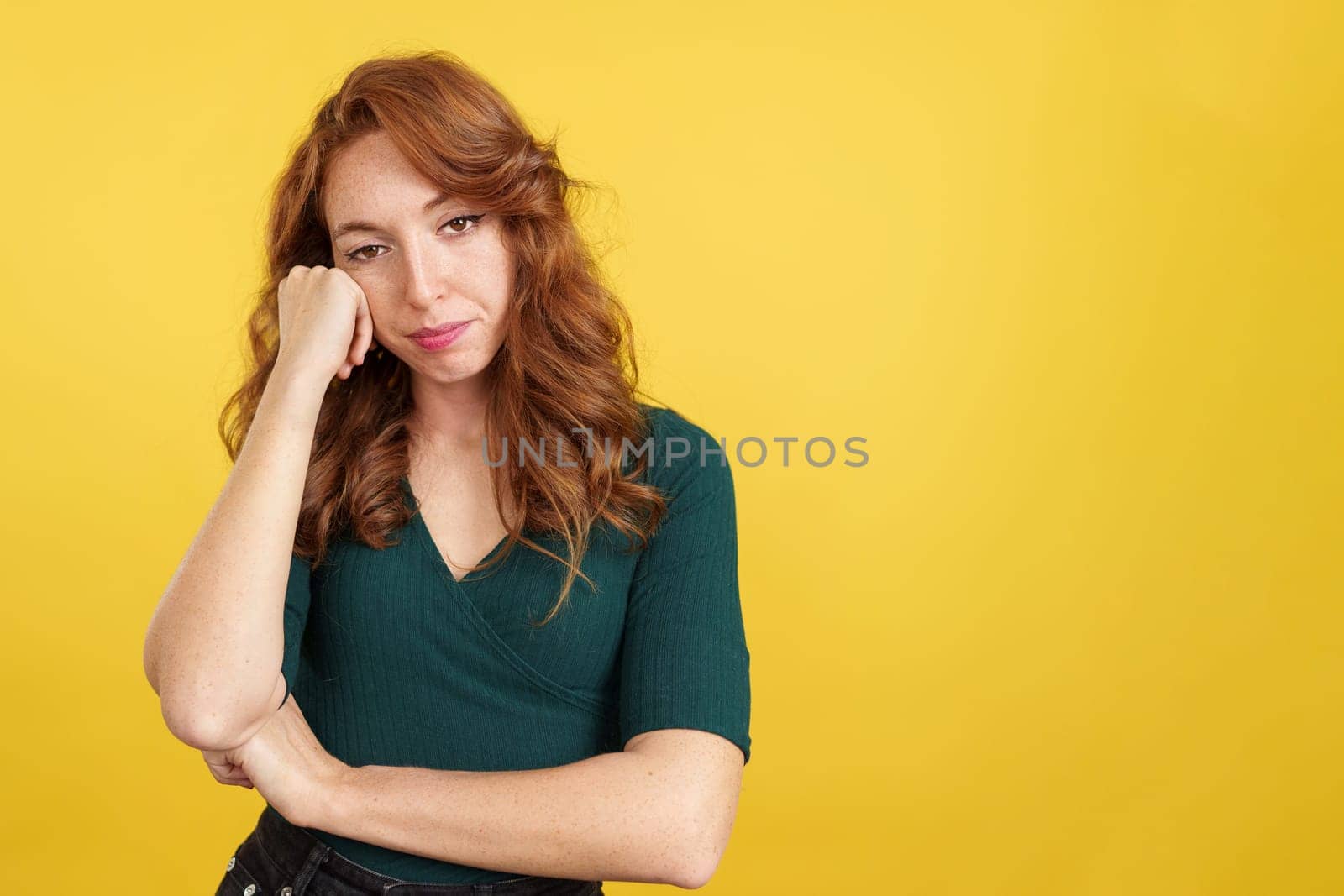 Bored redheaded woman standing and looking at the camera in studio with yellow background
