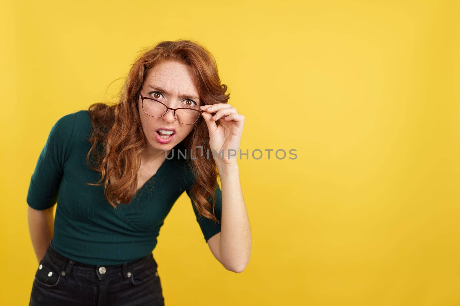 Redheaded woman with glasses staring at the camera in disbelief in studio with yellow background