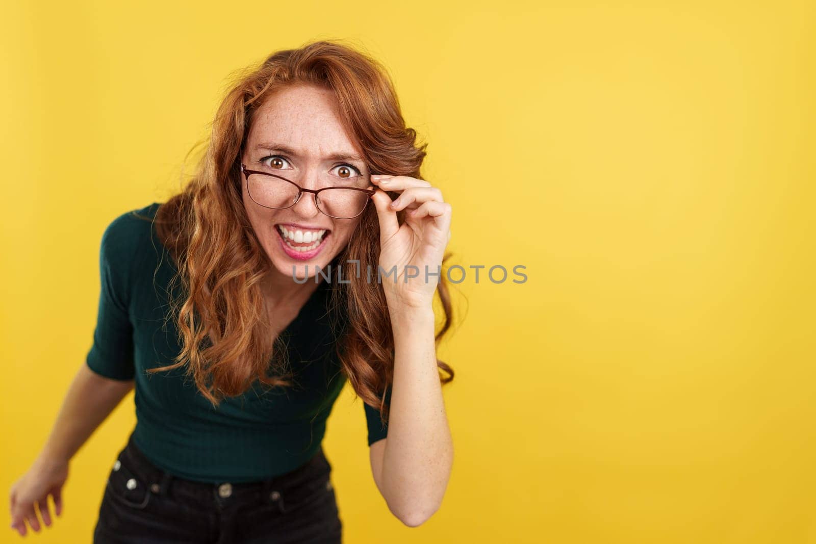 Redheaded woman with glasses looking at the camera with surprise in studio with yellow background