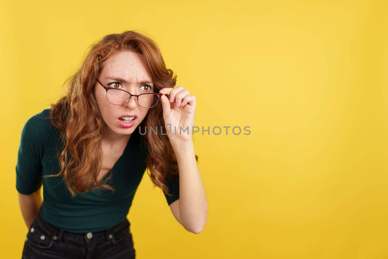 Redheaded woman with glasses spying with curiosity in studio with yellow background